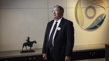 A man stands in a room looking off towards the left. Beside him is a small bronze statue and behind him is an art piece hanging on the wall.