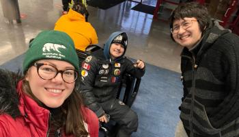 Three people bundled up for cold temperature are waiting in an airport. They smile at the camera.