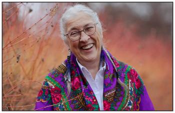 A woman stands in a field of long grasses. She has a colourful shawl over her shoulders. She smiles widely towards the camera.