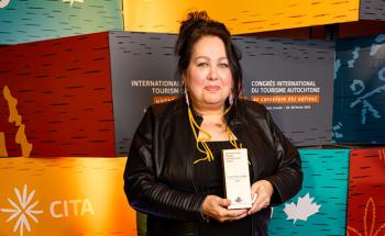 A woman stands in front of a background over blocks of colour. She is holding a trophy in front of her.