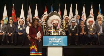 A woman and a man stand at a podium. The man wears a chief's feather 