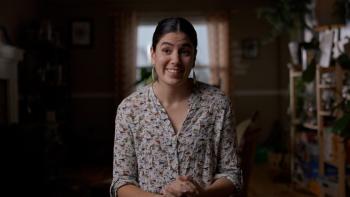 A smiling woman sits on a chair in a room that has plants around. Her hands folded in front of her.