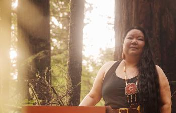 A woman stands in a forest. Sunbeams filter between the trees.