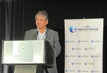 A man in a grey suit stands at a lectern. Beside him is a banner with a logo, a blue circle with a white feather in it, and writing that reads ICE Indigenous & Community Engagement 