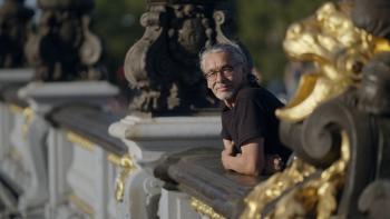 A man with grey hair and beard stands on a balcony. A gold gilded statute beside him.