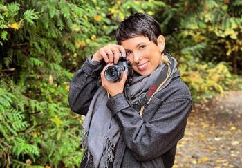 A young woman stands on a path amongst the trees. She smiles and has raised a camera up to her face.