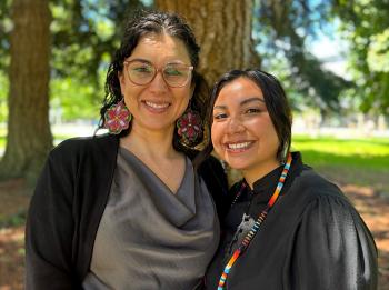 Two women stand among the trees in a park. They are close together and smile towards the camera.