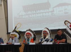 Chiefs in headdresses sit in front of a photo of a residential school