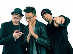 Three Indigenous men dressed in black jackets pose for the camera. One with his hand on his heart. Another with his hand in front of his chest in a prayer, and the other man pointing his finger towards the camera.