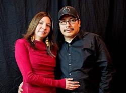 A woman in a red top stands with a man in a black shirt and ball cap, their arms around each other's waists.