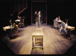 The character of a residential school student stands centre stage surrounded by students sitting at old-fashioned school desks.