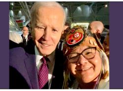 President Joe Biden takes a selfie with RoseAnne Archibald. He wears a blue suit and is smiling into the camera. She wears a birchbark headdress with a beaded medallion in the design of the AFN logo at her forehead.