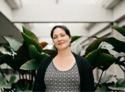 Behind a woman who stares toward the camera is a backdrop of large leafy indoor plants.