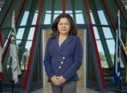 A woman stands in front of flags inside a building with a lot of windows. She is wearing a blue jacket, has her hands folded in front of her. She has dark shoulder-length hair.