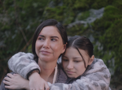 A young girl stands behind her mother wrapping her arms around the woman's shoulders.