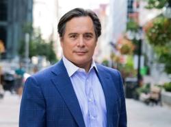 A man in a blue suit is on a tree covered city street. He looks at the camera.