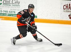 A hockey playing wearing a black uniform with orange accents has the puck on his stick.