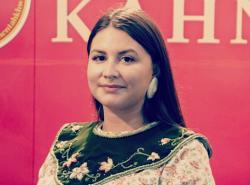 A woman in a traditional cotton dress with beaded collar stands against a red background smiling slightly.