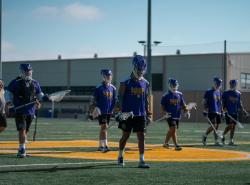 Lacrosse players walk across the playing field. They wear purple jerseys. 