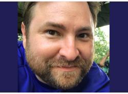 A man in a blue T-shirt looks directly into the camera. He has a beard and a mustache.