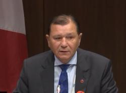 A man in a suit speaks. Behind him is a Canadian flag.