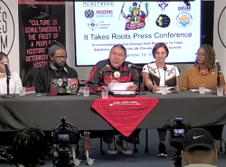 A group of people sit at a table with microphones on it. There are banners that read "The Peoples' Forum" on each end and behind the group.