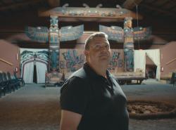 In the background are huge winged totem poles on each end of a painted cedar wall screen. Painted ceremonial curtains can be seen as are other cedar carved objects. A stepped wooden seating area flows upwards on either side of a dirt floor in the large building. In the centre is a place for a ceremonial fire. A man stands in the foreground looking upwards.