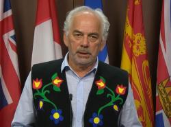 A man stands at a microphone in front of a row of flags.