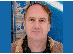 A man is photographed in front of a building with peeling blue paint on its walls.