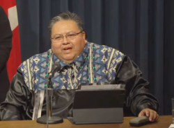 A man sits at a microphone and reads from notes set up in front of him He wears a black shiny ribbon shirt with a blue and white geometric design across the shoulders.