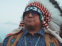 A man wearing a chief's feather bonnet looks off into the distance.