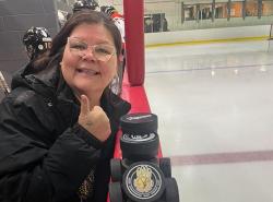 A woman stands in the players box in a ice rink. She raises her thumb as she smiles at the camera. A stack of pucks sit on top of the boards in front of her.