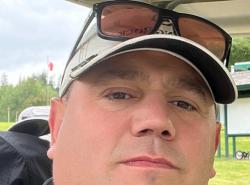 A man looks into the camera. He wears a cap with a pair of sunglasses perched on it.