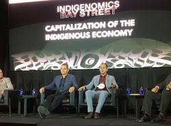 Four men sit on a stage in front of a wide screen on which is printed Capitalization of the Indigenous Economy over some art work in the West Coast style.