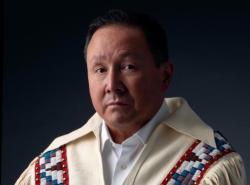 A man with a white fringed jacket with beading over the shoulders looks into the camera without smiling.