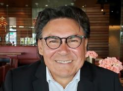 A man sits at a table in a restaurant. Flower arrangements are on tables. He smiles into the camera.