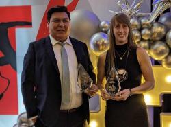 Two people, a man and a woman, are holding awards. In the background is a set made up of banners and balloons.