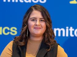 A young woman with long dark hair wears a mustard coloured top under a black vest. She stands in front of a blue screen with the word "mitacs" printed in white on it.