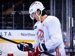 A man wearing a Washington Capitals hockey uniform is standing in the players' bench smiling and looking at the stick in his hands.