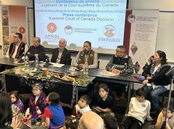 Children sit on the floor in front of a table with cultural items on it and behind that sits at a table First Nations leaders.