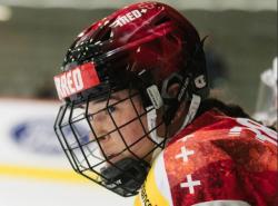 A young woman looks toward the ice. She wears a red hockey helmet with face cage.