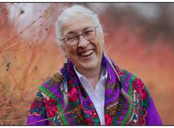 A woman stands in a field of long grasses. She has a colourful shawl over her shoulders. She smiles widely towards the camera.