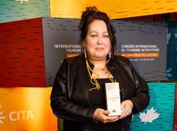 A woman stands in front of a background over blocks of colour. She is holding a trophy in front of her.