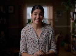 A smiling woman sits on a chair in a room that has plants around. Her hands folded in front of her.