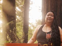 A woman stands in a forest. Sunbeams filter between the trees.