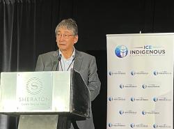 A man in a grey suit stands at a lectern. Beside him is a banner with a logo, a blue circle with a white feather in it, and writing that reads ICE Indigenous & Community Engagement 