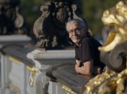 A man with grey hair and beard stands on a balcony. A gold gilded statute beside him.