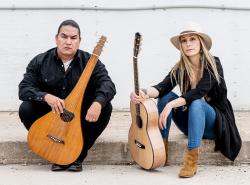 A man and a woman sit on a cement step in front of a white painted wall. Each hold a guitar. The man's guitar is a lap steel with a blunted headstock and a body that is round at the bottom but tapers up the neck.