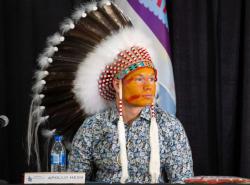 A young man wears a full feather bonnet and ocher-coloured face paint. He sits near a microphone and his name appears on a placecard in front of him--Apollo Hess.  