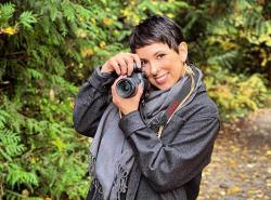 A young woman stands on a path amongst the trees. She smiles and has raised a camera up to her face.